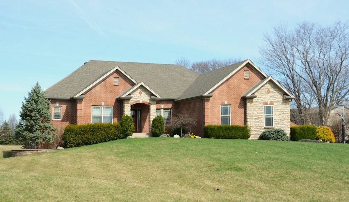A brick and stone ranch house with a shingle roof above a sloping lawn